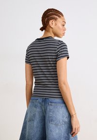 Μαύρο και άσπρο ριγέ T-shirt με κοντά μανίκια, στρογγυλή λαιμόκοψη, εφαρμοστό σχέδιο, σε συνδυασμό με ανοιχτό μπλε τζιν σορτς, δείχνοντας πίσω πλευρά.