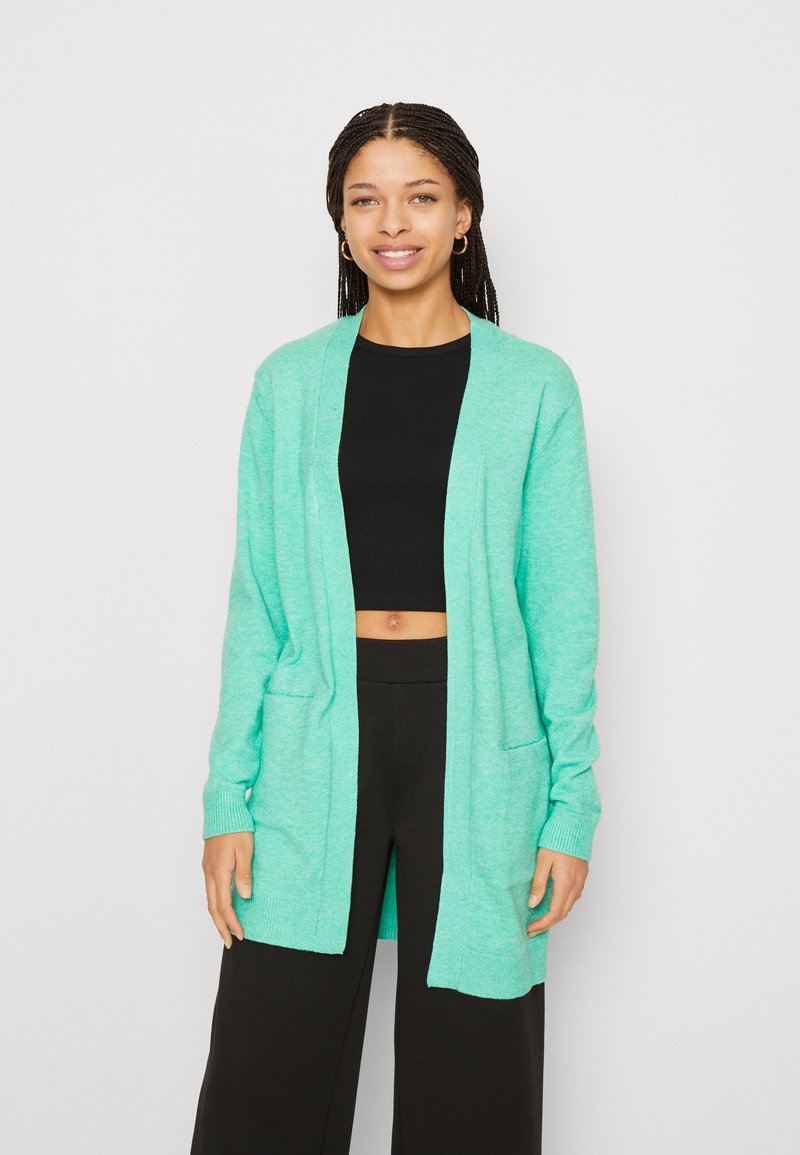 Gilet en maille vert menthe avec des manches longues, un devant ouvert, deux poches et des bords côtelés. Associe à un crop top noir et un pantalon.