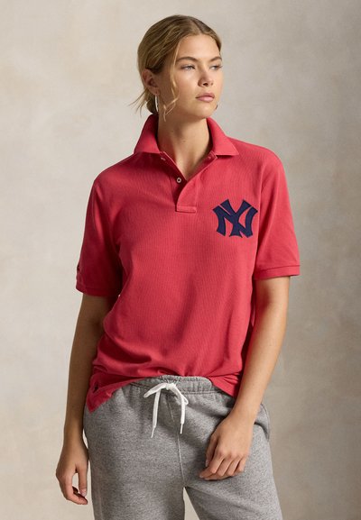 Νεαρή γυναίκα με κόκκινο πόλο μπλουζάκι με μπλε σκούρο λογότυπο και γκρι φόρμες, στέκεται μπροστά σε ουδέτερο φόντο, κοιτάζοντας προς τα δεξιά.