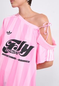 Rosa oversized skjorta med vertikala ränder, med en off-shoulder design. Svart "Fly Tanzbar" text och Adidas tre-rands accenter.