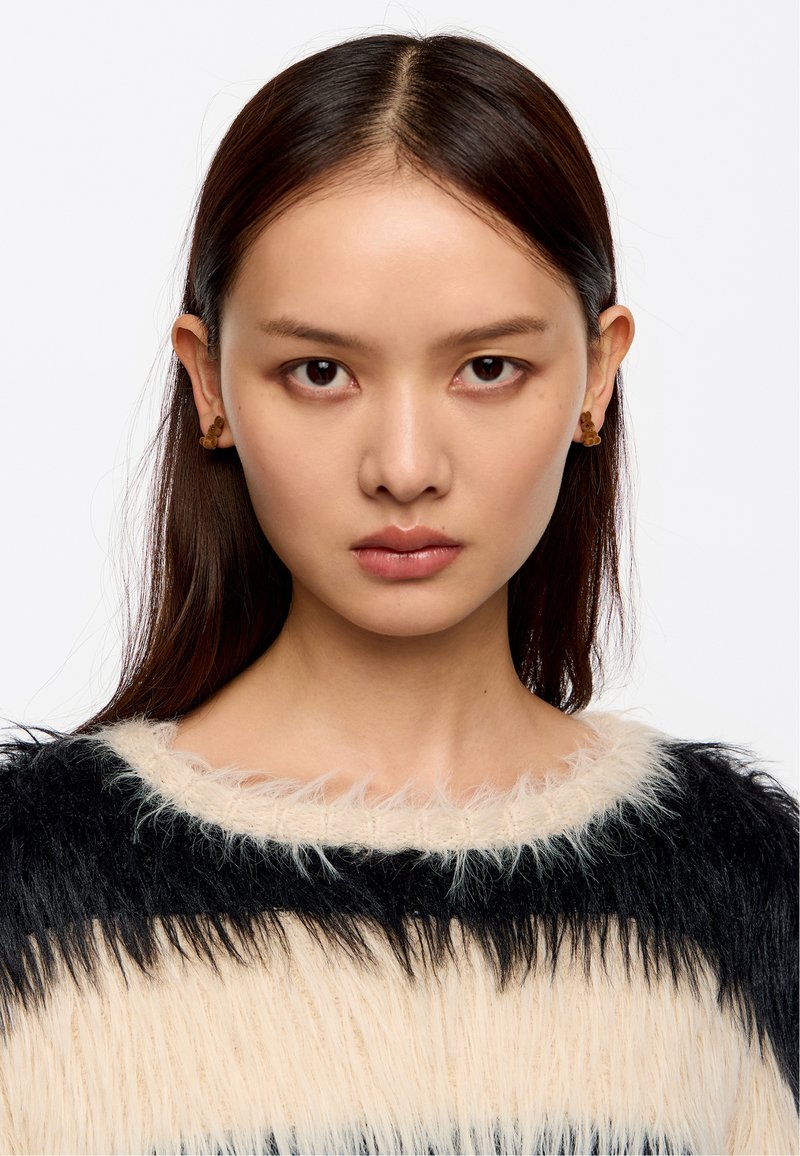 Modelo con largo cabello oscuro que lleva un suéter de pelo, a rayas en crema y negro. Pendientes redondos marrones notables contrastan con la textura.