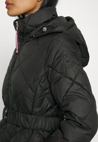 Black quilted jacket featuring a hood, elastic waistband, and contrasting zipper detail. Circular brand logo on the left sleeve.