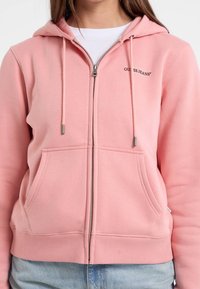 Felpa rosa con zip realizzata in tessuto morbido, caratterizzata da una tasca a marsupio, lacci e logo sul petto. Polsini e orlo aderenti.