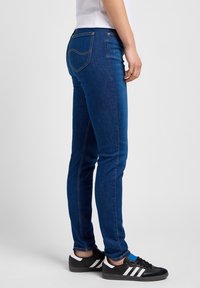 Smala blå denimjeans med kontrasterande orange sömmar, två bakfickor och midjehög midja. Bärs med svarta sneakers.