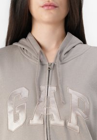 Ung kvinne iført en lys grå hettejakke med satin "GAP"-bokstaver, hettetråder synlige, utsnitt fra nese til øvre bryst.