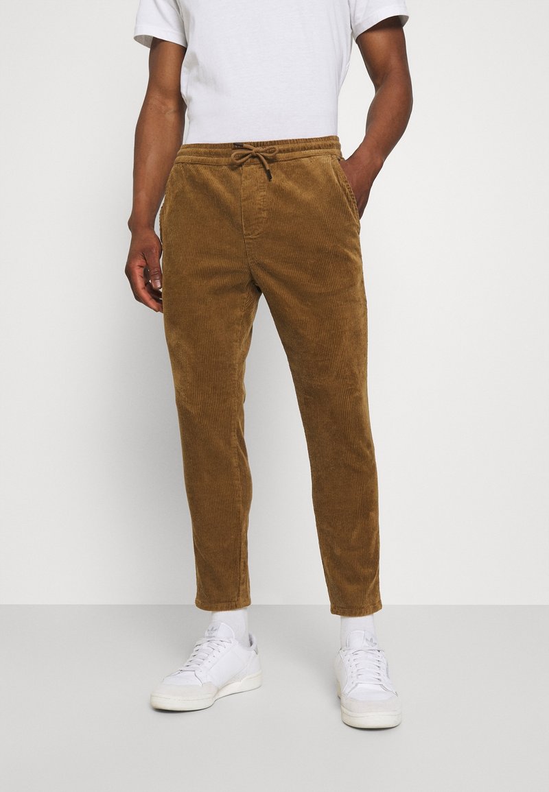 Man in bruine corduroy broeken met een trekkoord, witte sokken, witte sneakers en een wit shirt met korte mouwen, staande tegen een effen achtergrond.