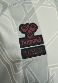 Ljust grått tyg med ett geometriskt mönster. Har en svart och vinröd broderad logotyp med texten "hummel HALO" under.