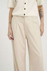 Beige vida byxor i lätt tyg med framfällor och en mjuk textur. Stylad med en stickad vit topp och svarta knappar.