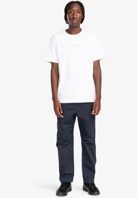 Camiseta de algodón blanca con un pequeño logo en el centro, combinada con pantalones azul marino y zapatos negros. Diseño simple, corte relajado.