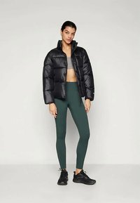 Svart dunjakke med høy krage, blank tekstur, kort design. Kombinert med grønne leggings og svarte joggesko.