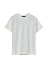 Camiseta de manga corta en un tono beige claro, fabricada con algodón suave. Presenta un cuello redondo y un pequeño parche de logo en la esquina inferior izquierda.