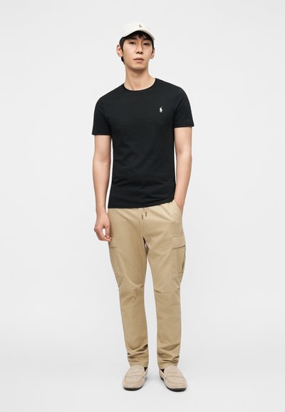 Μαύρο βαμβακερό μπλουζάκι με λογότυπο, ταμπά παντελόνι cargo με πλαϊνές τσέπες, μπεζ παπούτσια τύπου slip-on και ανοιχτόχρωμο καπέλο.