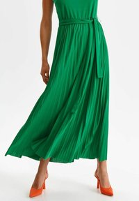 Grön plisserad maxiklänning med ett matchande bälte, som har en slät textur. Bärs med klarröda högklackade skor.