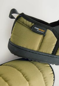 Zelené slip-on topánky z materiálu Cordura s čiernou textúrovanou gumovou podrážkou, polstrovaným dizajnom a štítkom s nápisom "Cordura" v bielej a oranžovej farbe.