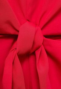 Red fabric with a smooth texture, featuring a knotted detail at the center and flowing ribbon-like ties extending from the knot.