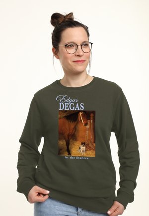Frau trägt ein olivgrünes Sweatshirt mit dem Gemälde "Bei den Ställen" von Edgar Degas, das ein Pferd und einen Hund in einem Stall zeigt.