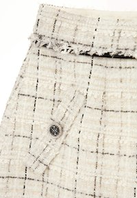Tissu en tweed blanc avec un motif en grille, présentant des bords effilochés. Un bouton noir avec une bordure en argent embellit la zone de la poche.
