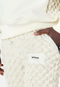 Witte fleece hoodie en gekreukte crèmekleurige joggers. Joggers hebben een ritszak en een genaaid label met "SIXTH JUNE PARIS." Kenmerkend geometrisch patroon.