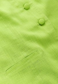 Close-up van limoengroen stof met geweven textuur, twee stoffen beklede ronde knopen en een horizontale bieszak.