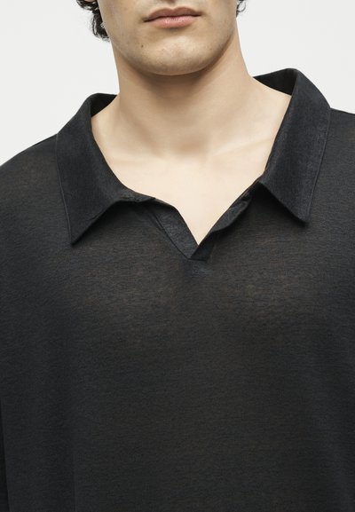 Νέος ενήλικας που φοράει μαύρο πουκάμισο με γιακά και V-σχήματος ντεκολτέ, εμφανιζόμενος από τους ώμους έως το στόμα σε απλό φόντο.