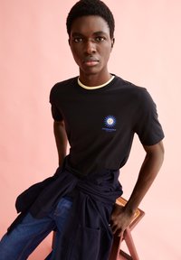 T-shirt in cotone nero con scollo rotondo e bordatura gialla, decorata con un logo blu. Il modello indossa jeans blu con una cintura scura.