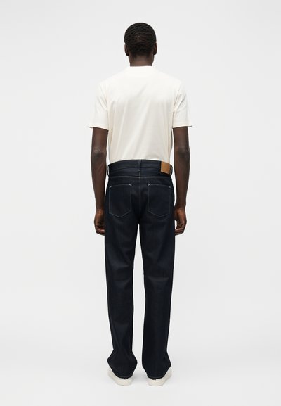 Mees seisab vaataja poole seljaga, kandes lihtsat valget lühikeste varrukatega särk, tumelõhitud teksad ja valged vabaajajalatsid valgel taustal.