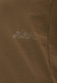 Débardeur marron en tissu lisse, avec des imprimés blancs pour la marque et les instructions d'entretien. Design simple sans embellissements supplémentaires.