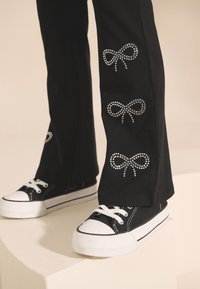 Melnas bikses ar rhinestone bantīšu dizainu ap apakšu, kombinētas ar melnām augstām sneakerēm un baltām gumijas zolēm.