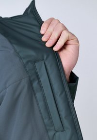 Donkergrijze zip-up jas met een zachte, glanzende textuur. Voorzien van een ritszak en contrasterende stof op de klep. Hand houdt de rits vast.
