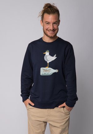 Navy blue sweatshirt featuring a graphic of a seagull in a hat standing atop a bottle, with a paper boat inside the bottle design.