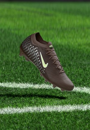 Gheată de fotbal maro cu logo alb și părți laterale texturate, plutind deasupra unui teren de iarbă verde, aproape de linia albă de delimitare.