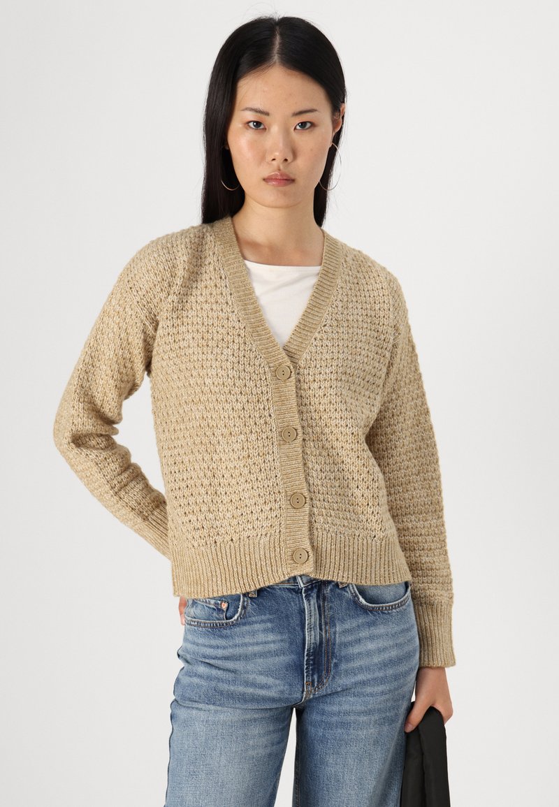 Cardigan en tricot beige avec un col en V, des manches longues et cinq boutons. Tissu texturé avec des poignets et un ourlet côtelés. Porté par-dessus une chemise blanche.