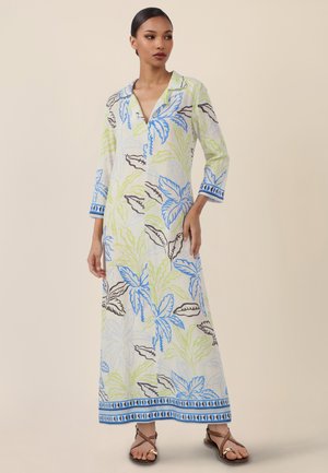 Frau trägt ein langes weißes Kleid mit blau-grün-braunen Blattmustern, dreiviertellangen Ärmeln und braunen, geschnürten Sandalen.