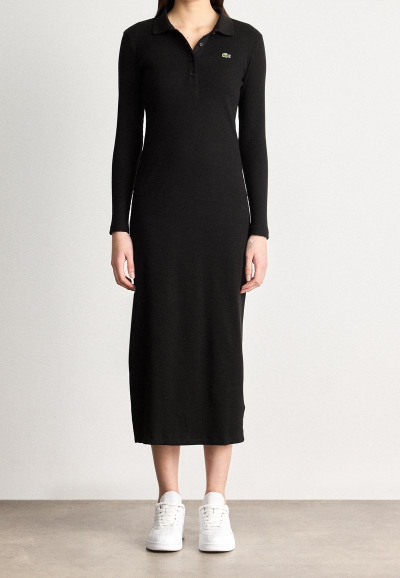 Femme portant une longue robe noire col avec manches longues et des baskets blanches, debout sur un fond clair uni.