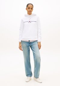 Biała bluza z kapturem z logo "TOMMY HILFIGER", sznurkiem i ściągaczem u dołu, zestawiona z szerokimi jasnoniebieskimi dżinsami i białymi trampkami.