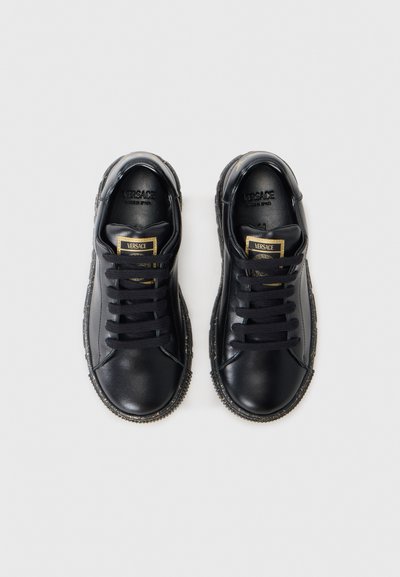 Zapatillas de cuero negro con punta redonda, cordones planos, detalles dorados y suelas de goma texturizada. Presentan una etiqueta de marca en la lengüeta.