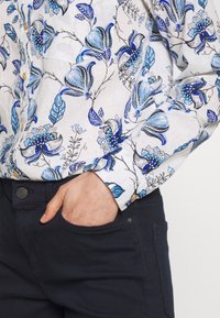 Primer plano de una persona que lleva una camisa blanca con patrones florales azules y pantalones oscuros, con la mano de manera casual en el bolsillo delantero.