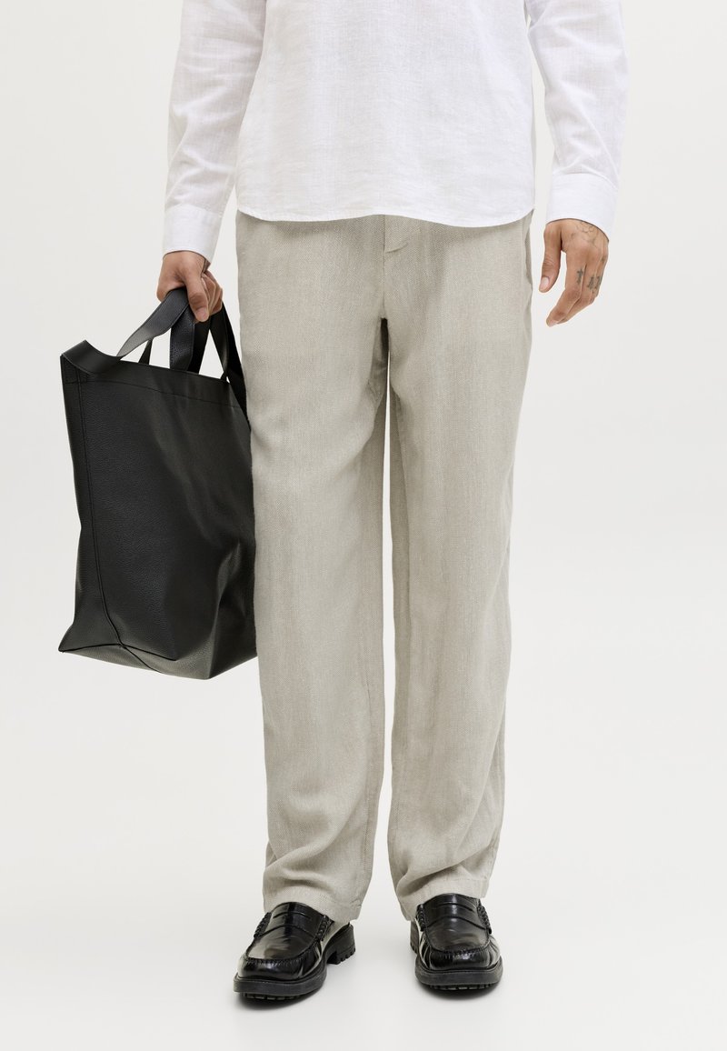 Άτομο που φοράει ανοιχτό μπεζ παντελόνι, μαύρα μοκασίνια και λευκό πουκάμισο, κρατώντας μια μεγάλη μαύρη δερμάτινη τσάντα ώμου στο αριστερό χέρι.