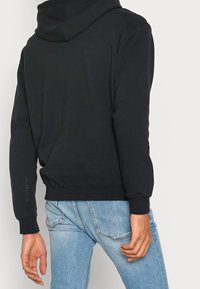 Svart huvtröja med mjuk textur, designad med en avslappnad passform, ribbade ärmslut och en logga på ärmen. Matchad med blå jeans.