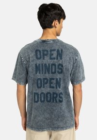 Lichtblauw denim T-shirt met een vervaagde textuur, met in het zwart gedrukte tekst: "OPEN MINDS OPEN DOORS" op de achterkant. Korte mouwen, ronde hals.