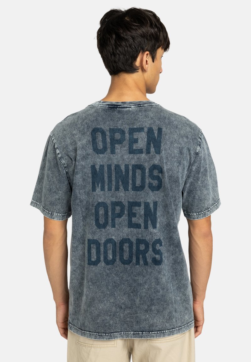 Lichtblauw denim T-shirt met een vervaagde textuur, met in het zwart gedrukte tekst: "OPEN MINDS OPEN DOORS" op de achterkant. Korte mouwen, ronde hals.