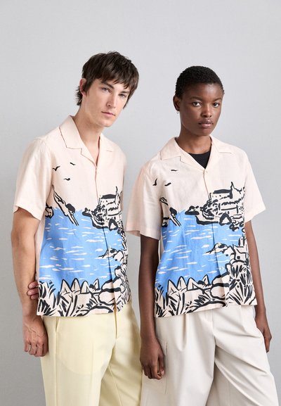 Chemises à manches courtes en rose clair avec un imprimé de scène de plage bleue ; présente des contours et des détails noirs ; coupe décontractée ; portée avec un pantalon beige et crème.