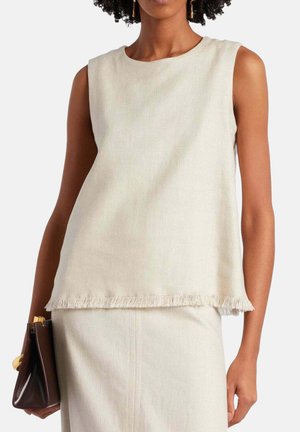 Femme portant un haut beige sans manches avec un ourlet à franges et une jupe assortie, tenant un sac à main marron foncé avec des détails dorés.