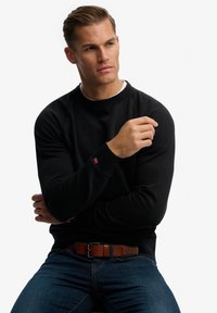 Felpa nera con colletto e polsini a coste, caratterizzata da un tessuto testurizzato. Indossata con jeans in denim blu e una cintura in pelle marrone.