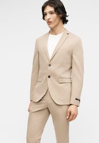 Νέος άνδρας με μπεζ κοστούμι δύο κουμπιών και αντίστοιχο παντελόνι, φορώντας λευκό μπλουζάκι με στρογγυλή λαιμόκοψη, στέκεται μπροστά σε ένα απλό λευκό φόντο.