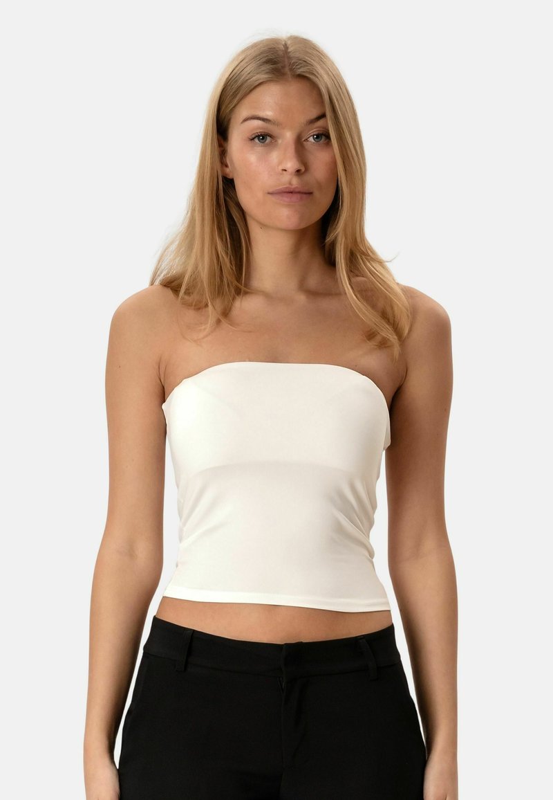Jeune femme aux longs cheveux blonds portant un crop top blanc sans bretelles et un pantalon noir, debout devant un fond clair uni.