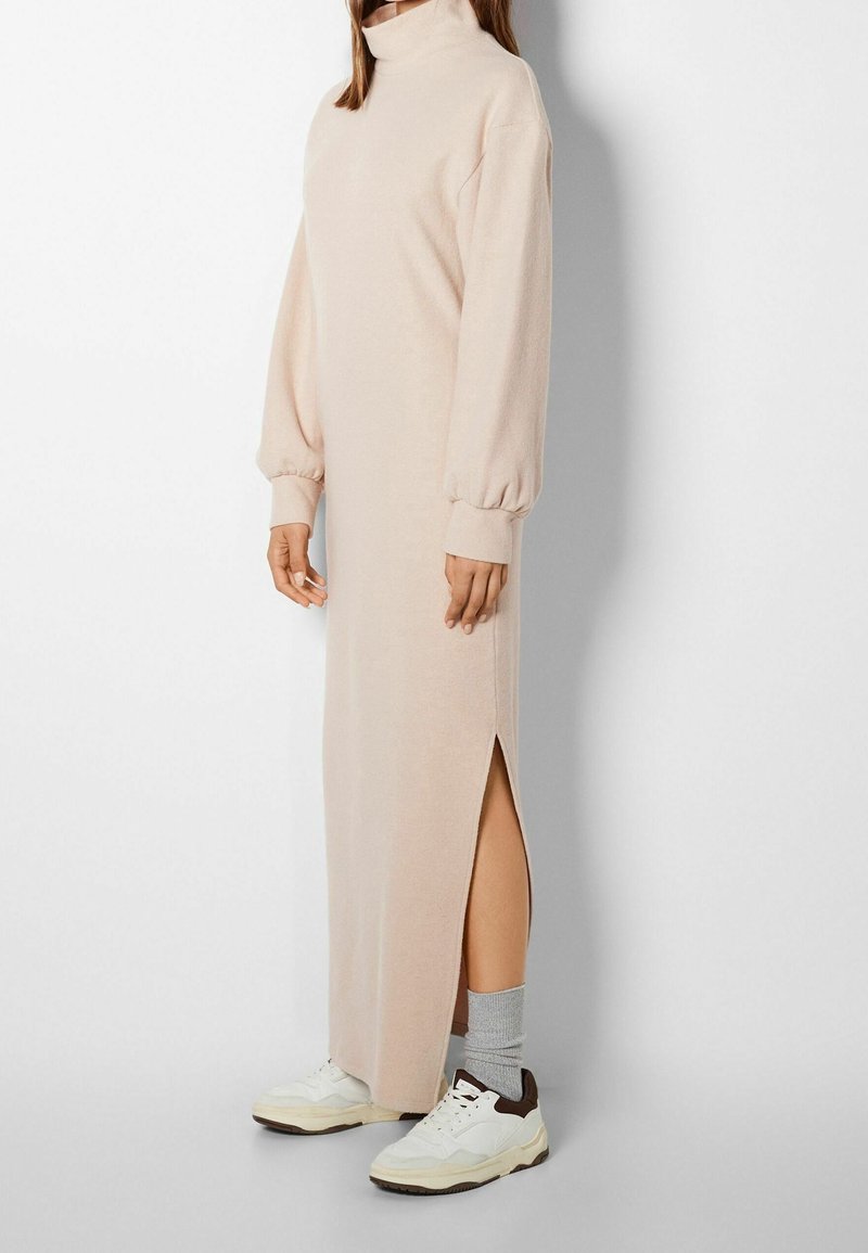 Robe longue, rose pâle, avec un col montant, des manches longues, une fente sur le côté et une texture douce, accompagnée de baskets blanches et brunes.
