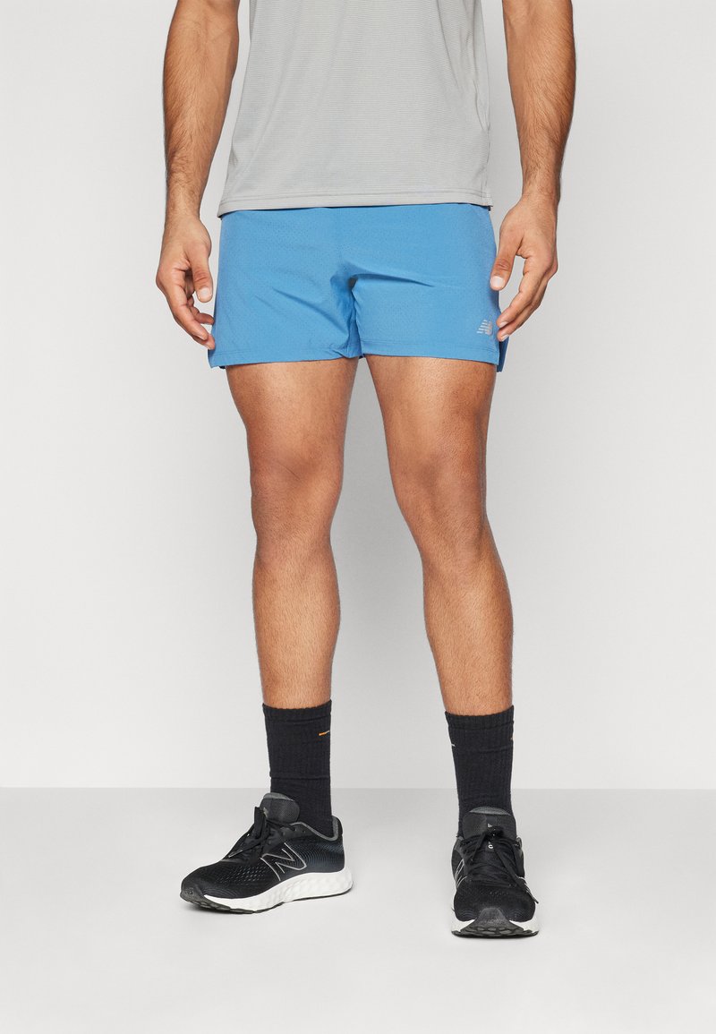 Calções de atletismo azuis para homem, tecido leve, cinturão elástico, combinados com sapatilhas de corrida pretas e meias de cano curto pretas. T-shirt cinza lisa.