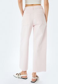 Pantalons en denim rose pâle à jambe large, dotés de deux poches arrière et d'un ourlet brut. Associés à des sandales de couleur claire.