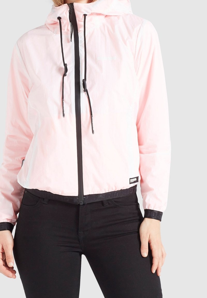 Jasnoróżowa kurtka przeciwwiatrowa z kapturem, czarnym zamkiem błyskawicznym i troczkami; posiada elastyczne mankiety i pas w talii z detalami logo.
